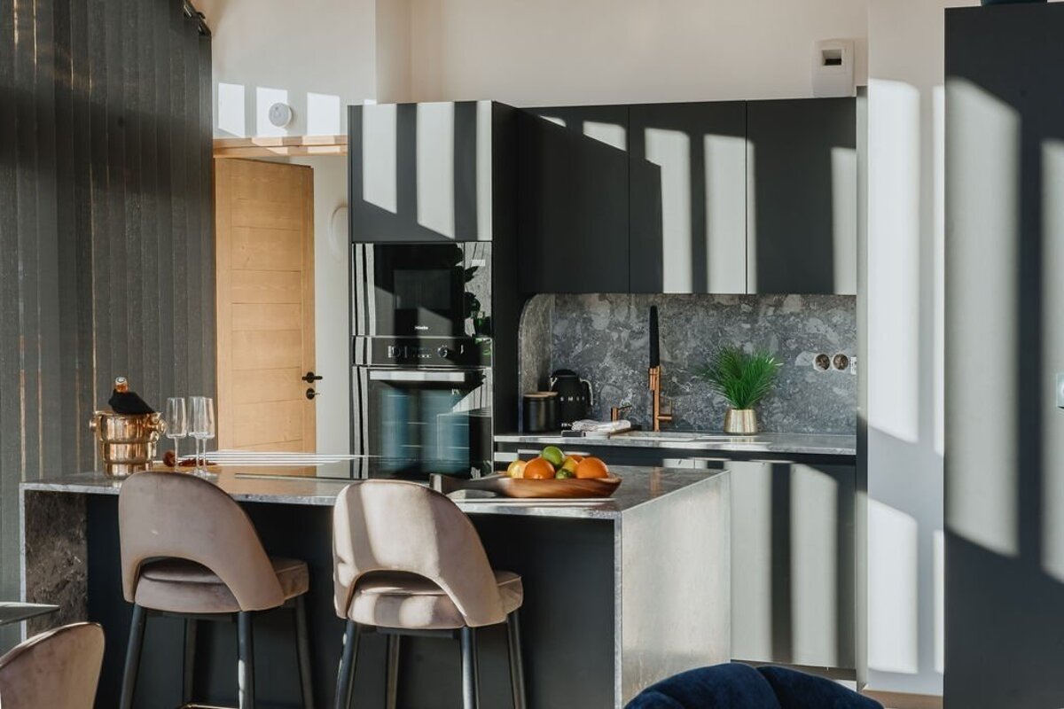 A sunlit modern kitchen with dark cabinetry, a stone island, and bar stools. Strong light casts geometric shadows across the room.