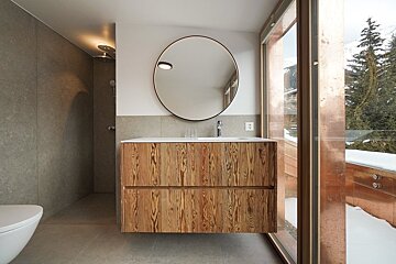 A sleek, modern bathroom with a wooden vanity, round mirror, and a large window overlooking a snowy mountain landscape.
