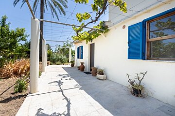 A sunny white house with vibrant blue shutters, partially covered by a vine-draped pergola, casting shadows on the patio.