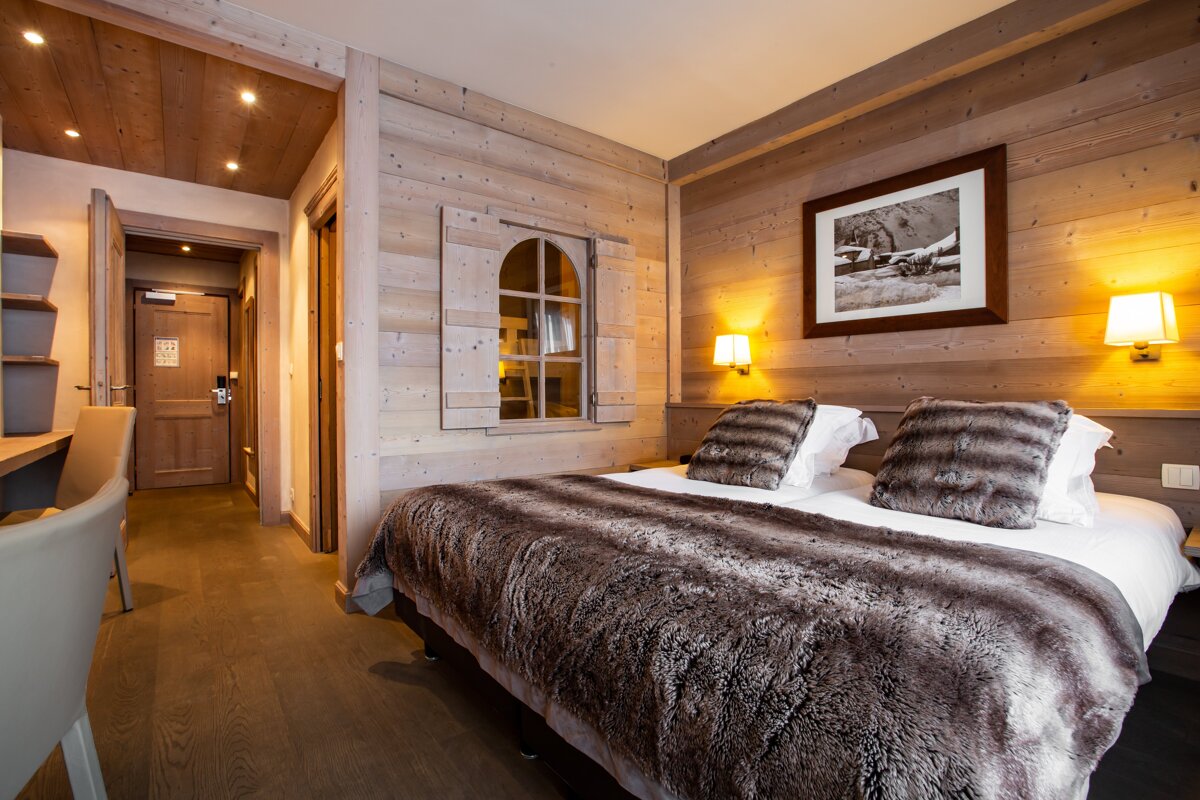 A cozy, wood-paneled chalet bedroom with a large bed, faux fur bedding, an arched window with shutters, and warm sconce lighting.