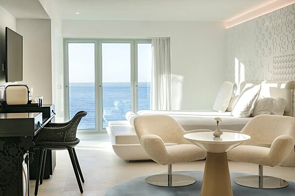 Bright, modern white hotel room with stunning ocean views, minimalist decor, and chic seating, bathed in natural light.
