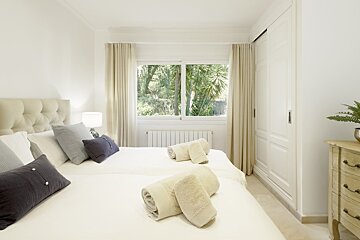 A bright, neutral bedroom featuring beds with rolled towels, grey pillows, and a large window looking out onto green trees.