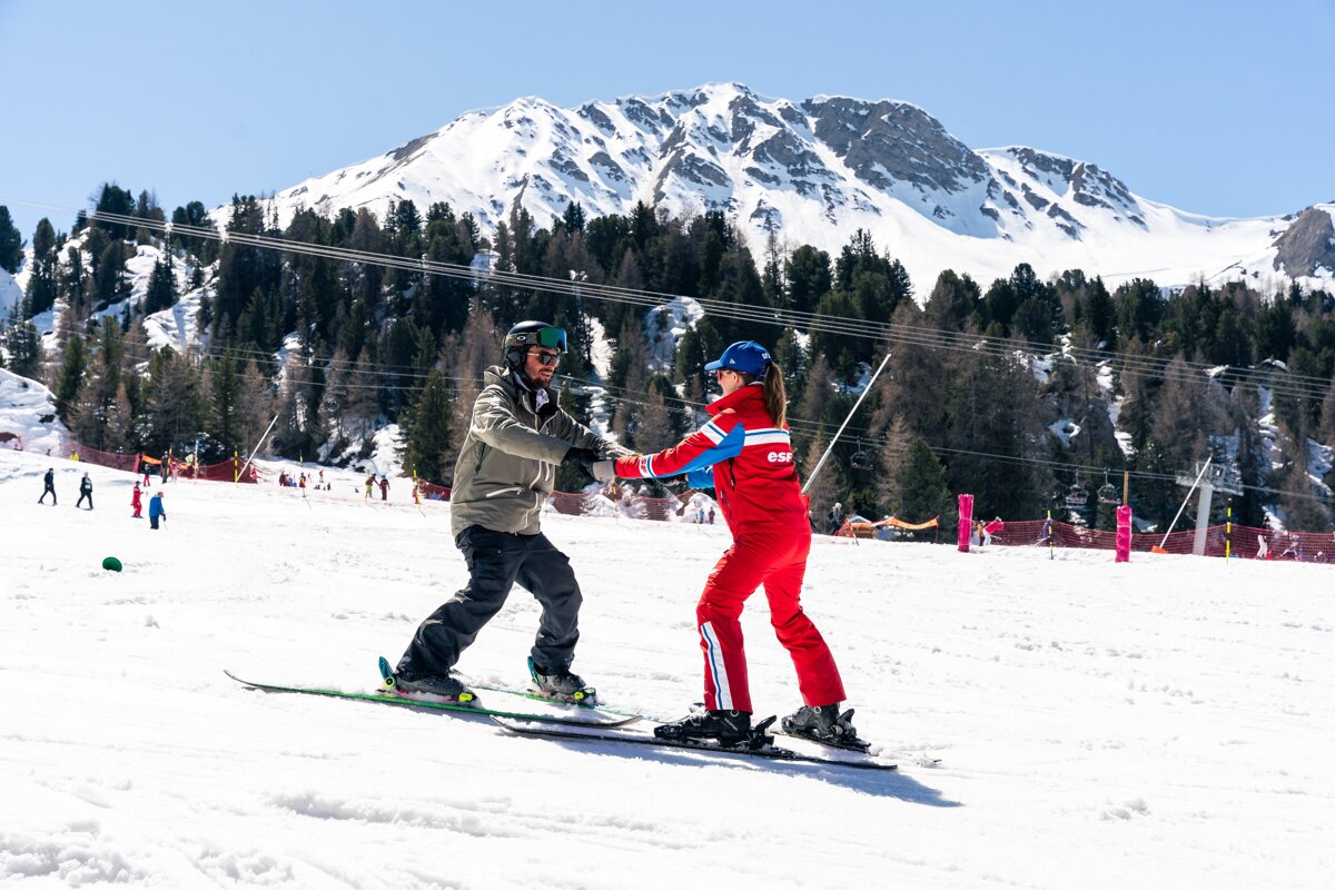 A ski instructor in red teaches a student on a sunny, snow-covered mountain, holding hands for support. Pine trees and snowy peaks are visible.