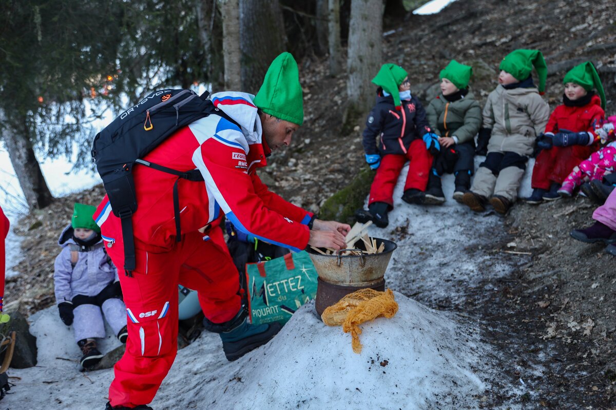 An adult in a red ski suit and green elf hat prepares a fire for children in green elf hats, gathered on a snowy hillside.