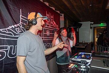 Two energetic DJs, a man in headphones and a woman gesturing, perform at a lively club event with DJ equipment and a graphic banner behind them.