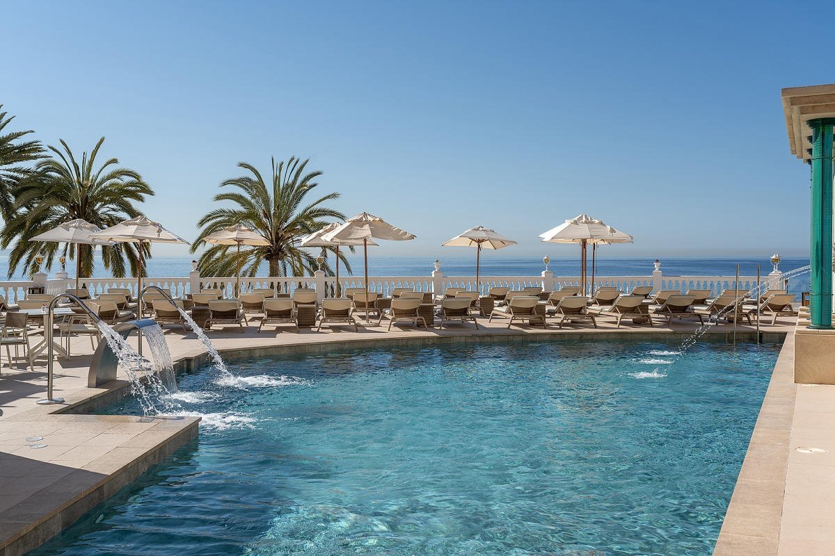 Sunny pool with waterfalls, palm trees, loungers, and umbrellas, overlooking the ocean under a clear blue sky.