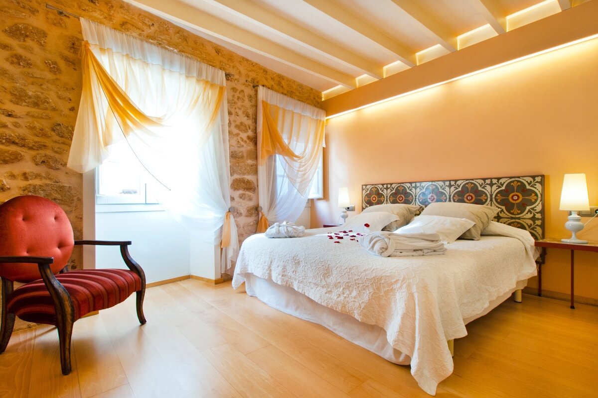 A cozy bedroom featuring stone walls, a large bed with a patterned headboard, rose petals, a red armchair, and warm ambient lighting.