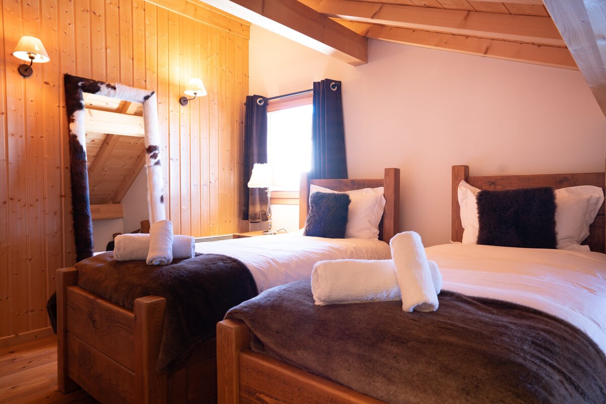 A cozy, rustic bedroom featuring two single wooden beds with white linens, dark throws, and rolled towels. A cowhide-framed mirror leans against the wood-paneled wall.