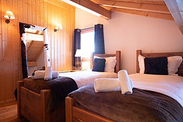 A cozy, rustic bedroom featuring two single wooden beds with white linens, dark throws, and rolled towels. A cowhide-framed mirror leans against the wood-paneled wall.