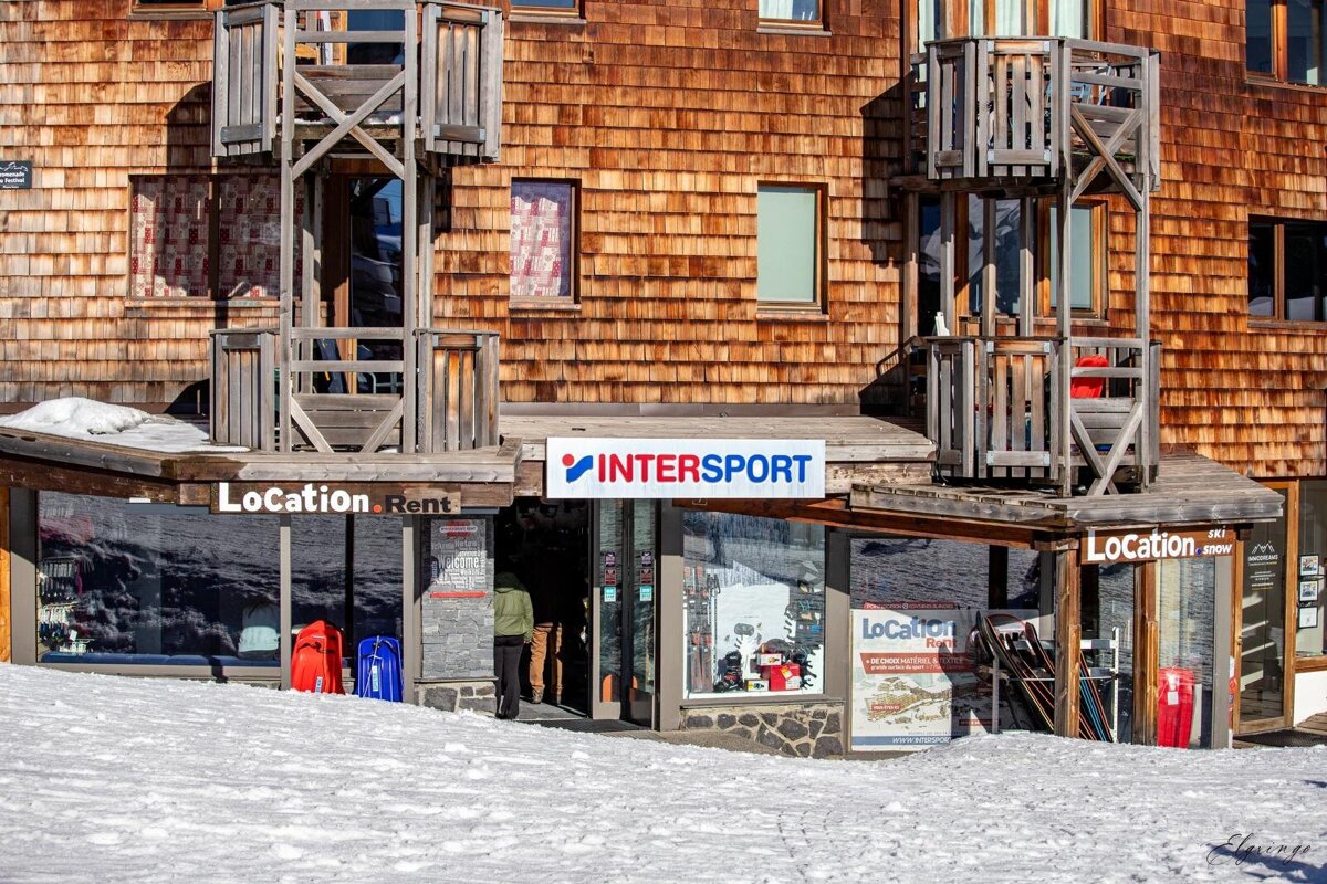 An Intersport ski rental shop with a rustic wooden shingle facade and balconies, surrounded by snow. A person is seen entering the store.
