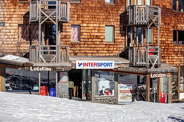An Intersport ski rental shop with a rustic wooden shingle facade and balconies, surrounded by snow. A person is seen entering the store.