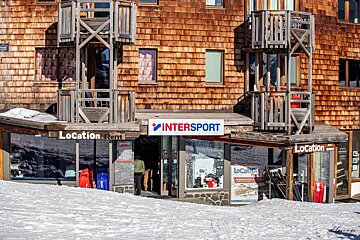 An Intersport ski rental shop with a rustic wooden shingle facade and balconies, surrounded by snow. A person is seen entering the store.