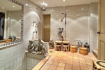A rustic, tiled bathroom with multiple showers, a bathtub adorned with statues, terracotta floor, wooden stools, and buckets.
