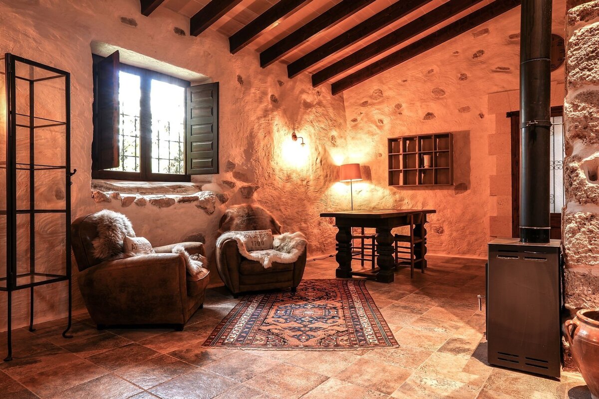 A warm, rustic room featuring textured stone walls, wooden beams, two cozy armchairs with fur, a patterned rug, and a modern wood stove.