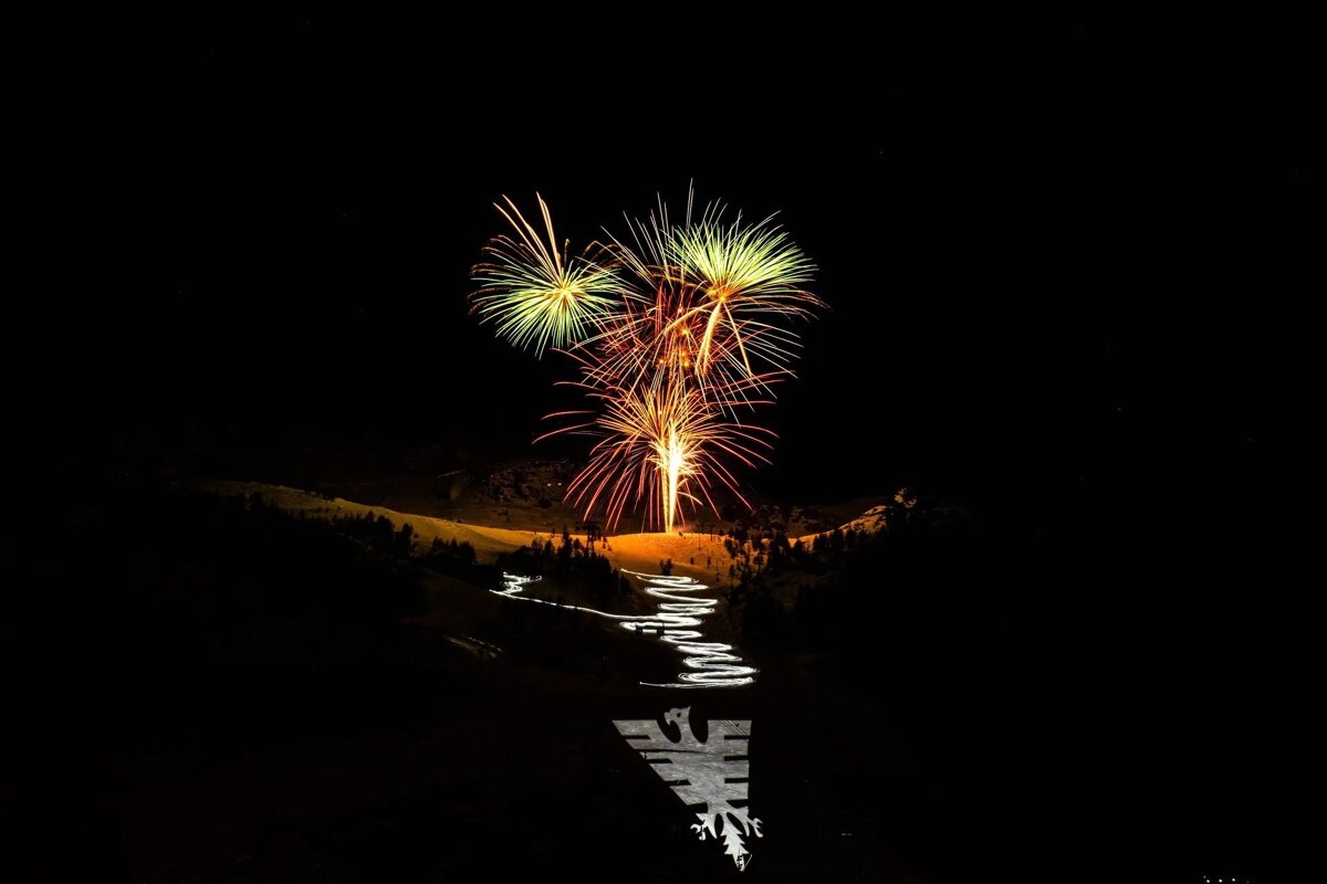 Bright fireworks explode above a dark, snowy mountain featuring a winding illuminated path and a large white snowflake design in the foreground.