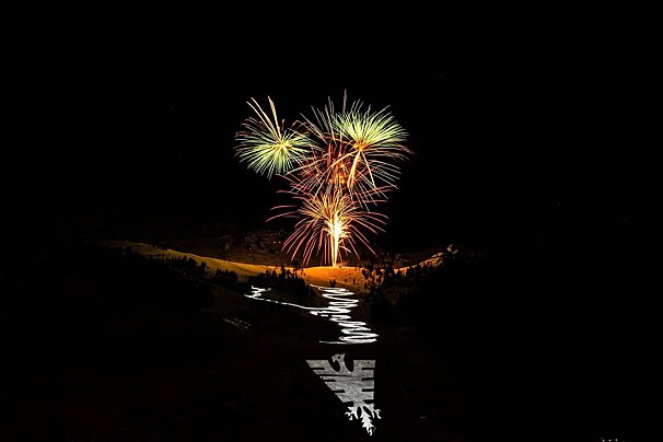 Bright fireworks explode above a dark, snowy mountain featuring a winding illuminated path and a large white snowflake design in the foreground.
