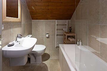 A cozy bathroom with a slanted wooden ceiling, beige tiled walls, a white sink, toilet, and bathtub. It features a towel warmer and wooden shelf.