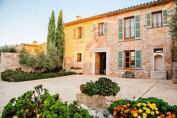 A beautiful stone house with green shutters bathed in warm sunlight, featuring a welcoming courtyard with vibrant flowers and tall cypress trees.