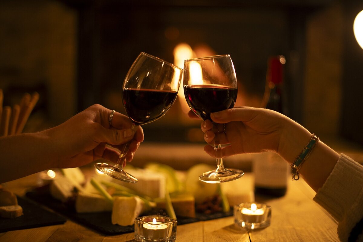 Two people toast red wine glasses by a warm fireplace. Candles, cheese, and snacks on the table create a cozy, romantic atmosphere.