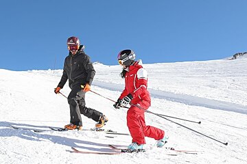Two skiers, a man and a woman, walk uphill on a snowy mountain under a clear blue sky. They are dressed in winter gear and helmets.