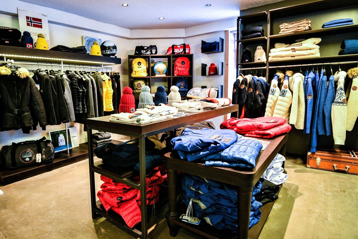 A cozy clothing store showcasing winter jackets, colorful hats, and bags on racks and display tables, with a Norwegian flag visible.
