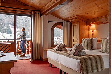 A man on a cozy, wood-paneled ski chalet balcony enjoys snowy mountain views from his rustic room, complete with a large bed.