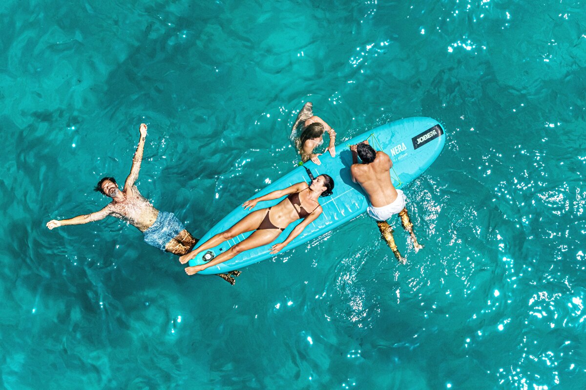 An aerial shot of four people relaxing and swimming in clear turquoise water with a blue paddleboard.