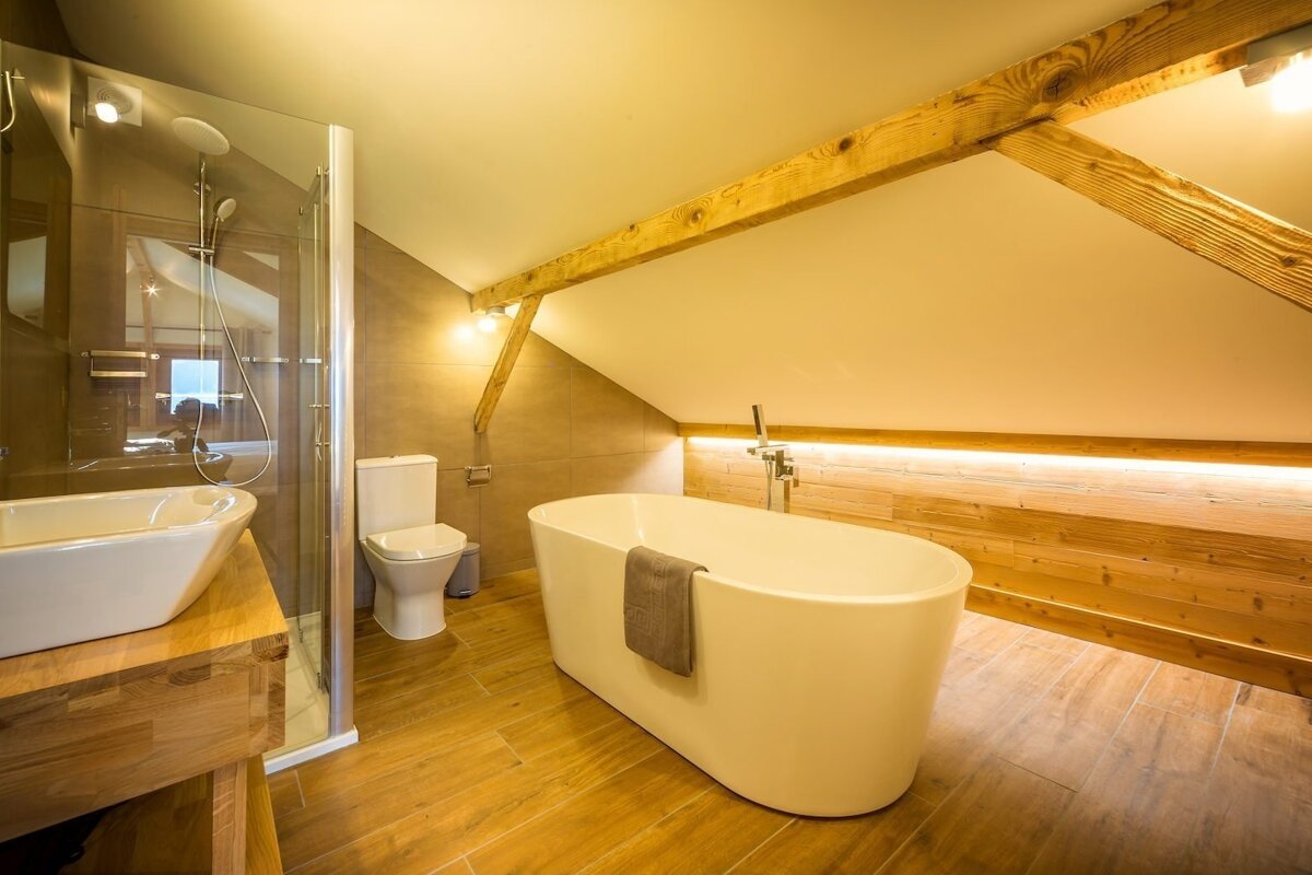A modern attic bathroom features a freestanding tub, walk-in shower, toilet, and warm wooden accents under a sloped ceiling with exposed beams.