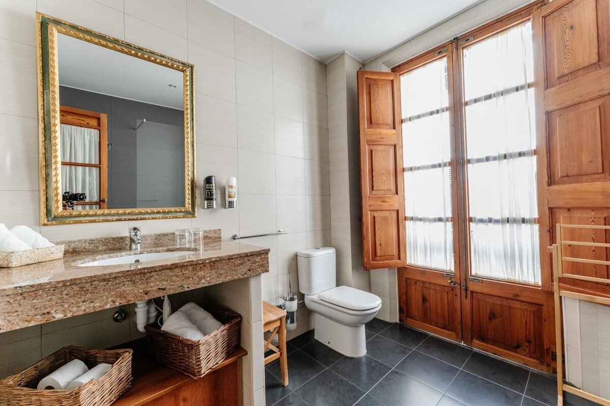 A bathroom with a granite counter, ornate mirror, toilet, and large windows with open wooden shutters. Baskets hold towels and supplies.