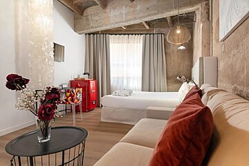 An industrial-chic room featuring a bed, beige couch, vibrant red mini-fridge, raw concrete ceiling, and a vase of dark red flowers.