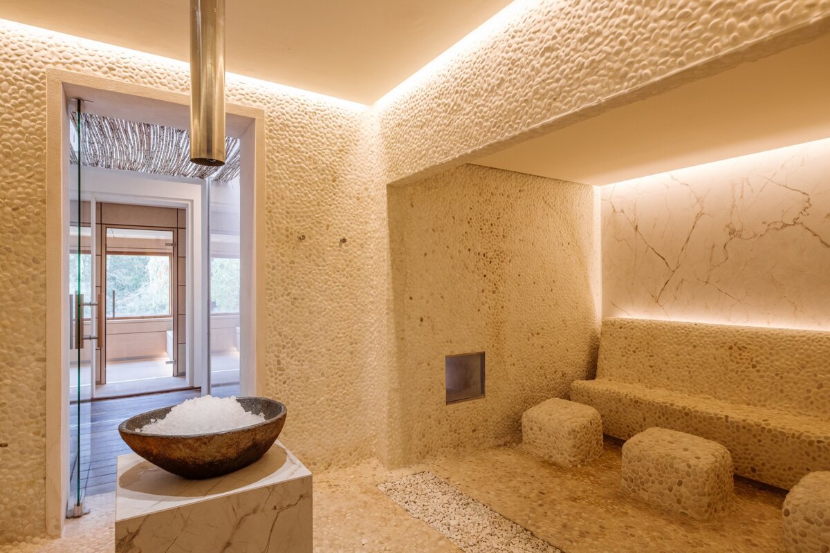 An elegant spa room with textured pebble walls, integrated warm lighting, stone seating, and a large bowl of ice for cold therapy.