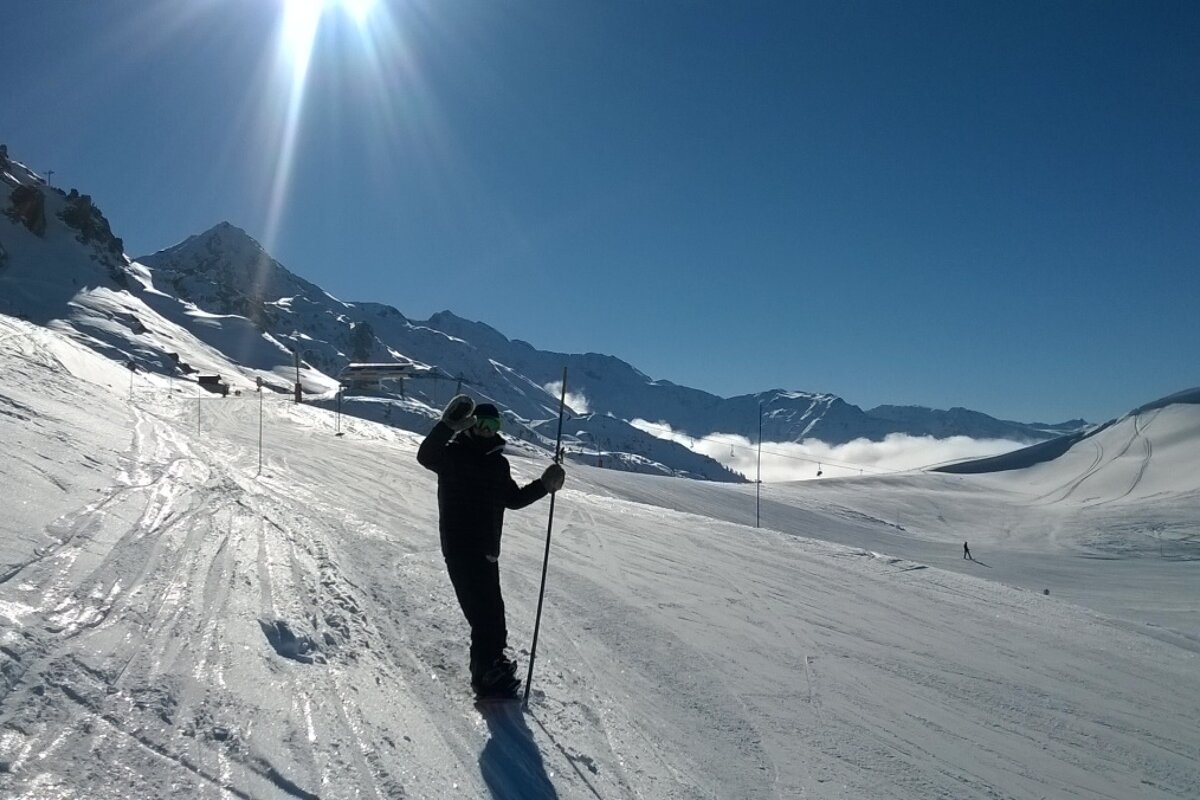 Snowboarder in the foreground holding onto a piste marker, briliant white snowy path with the vagere chairlift in the background and a very blue sky and sun