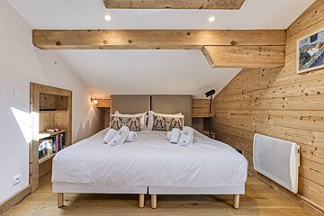 A cozy bedroom with a large bed, exposed wooden beams, and wood-paneled and white walls, creating a rustic yet modern feel.