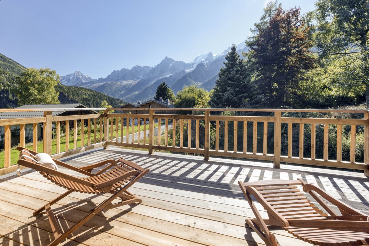 A sunny wooden deck with lounge chairs overlooks majestic snow-capped mountains, green forests, and a clear blue sky.