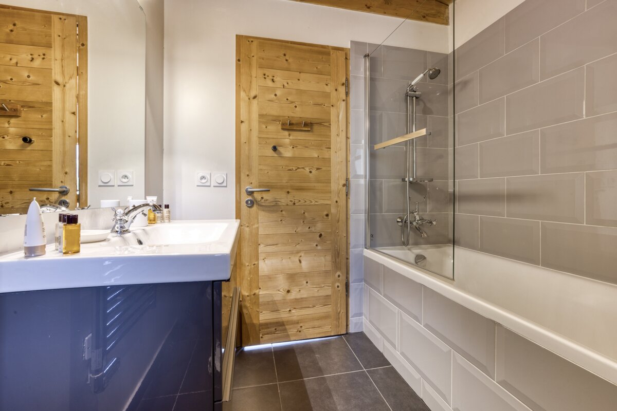 A modern bathroom featuring a white sink on a dark vanity, a wooden door, and a bathtub with a glass shower screen against grey tiled walls.