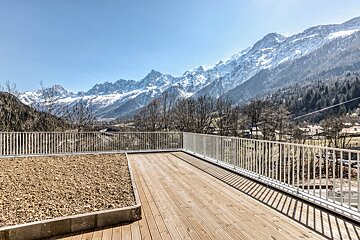 A large wooden deck with a white railing offers panoramic views of majestic snow-capped mountains, a valley, and a clear blue sky.