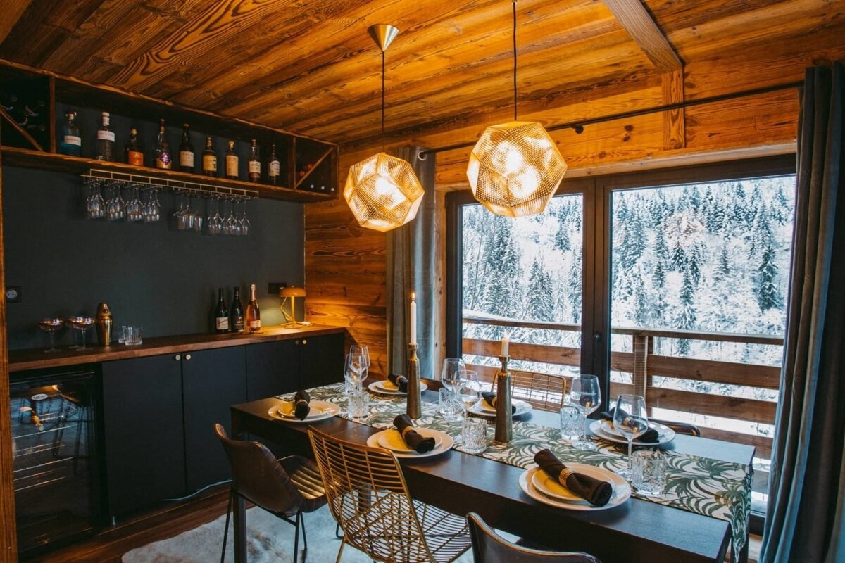 A warm chalet dining room features a set table, a bar, and large windows with a stunning view of a snowy winter forest. Rich wooden accents and modern lighting.