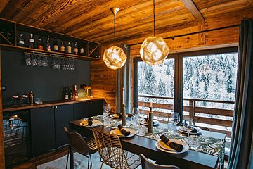 A warm chalet dining room features a set table, a bar, and large windows with a stunning view of a snowy winter forest. Rich wooden accents and modern lighting.