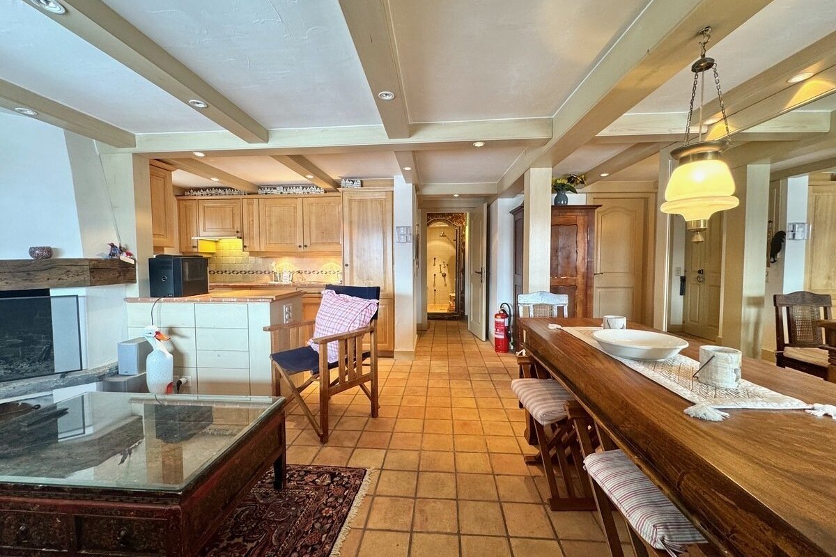 A spacious, rustic open-plan room with wood-beamed ceiling, tiled floor, featuring a kitchen, fireplace, and a long wooden dining table.