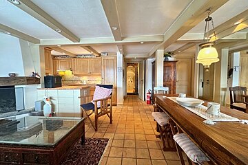 A spacious, rustic open-plan room with wood-beamed ceiling, tiled floor, featuring a kitchen, fireplace, and a long wooden dining table.