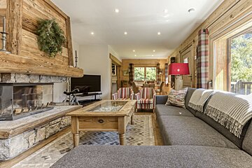 A cozy, rustic chalet living room features a stone fireplace, large gray sectional sofa, wooden coffee table, and striped armchairs. Wood paneling and a view of nature create a warm ambiance.
