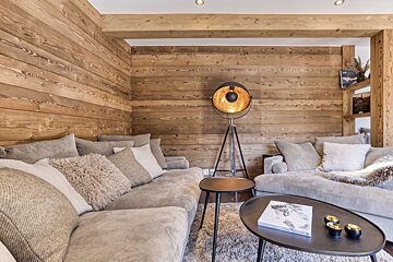 A cozy living room features rustic wood-paneled walls, plush light gray sectional sofas with numerous pillows, an industrial tripod floor lamp, and coffee tables on a textured rug.