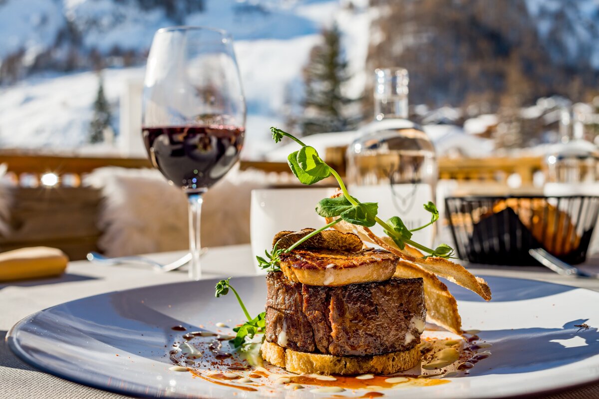 A gourmet steak and red wine are elegantly presented on an outdoor table, with a beautiful snowy mountain landscape in the background.