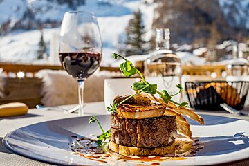 A gourmet steak and red wine are elegantly presented on an outdoor table, with a beautiful snowy mountain landscape in the background.