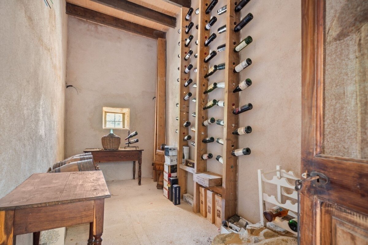 A rustic wine cellar with wine bottles displayed on vertical wooden racks, old wooden tables, wine barrels, and a small window.
