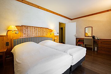 A cozy hotel room with two twin beds, wooden headboard, wall lamps, and a desk with a mirror and coffee machine. Features warm wood accents.