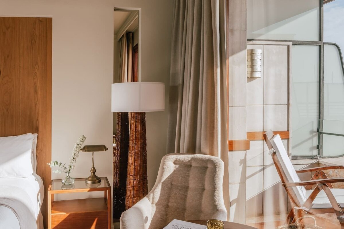 Sunlit room with a bed, elegant armchair, and floor lamp, featuring neutral tones and warm light spilling through the window.