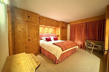 Cozy bedroom with light wooden built-in furniture around a bed with red patterned bedding, terracotta curtains, and a small desk.