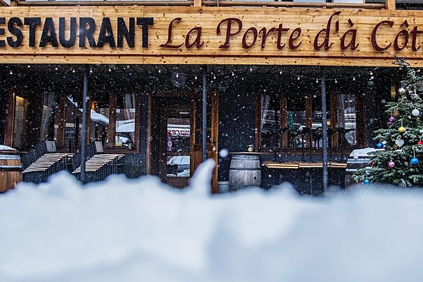 Snow falls outside a French restaurant, 'La Porte d'à Côté', decorated for Christmas with a tree and stacked outdoor seating.