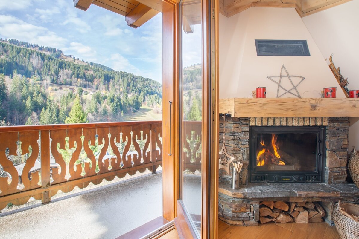 Cozy chalet interior with a burning fireplace, opening to a balcony with a stunning view of a forested mountain landscape.